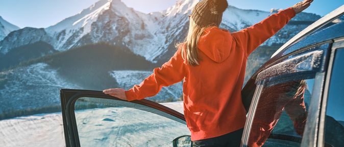 Eine Frau lehnt aus der offenen Autotür und schaut auf Schneebedeckte Berge
