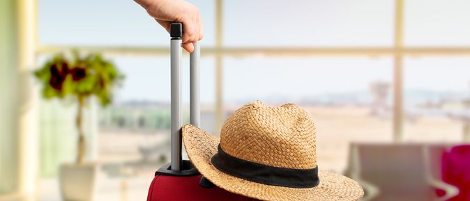 Eine Hand schiebt einen Roten Koffer auf dem ein Sonnenhut liegt