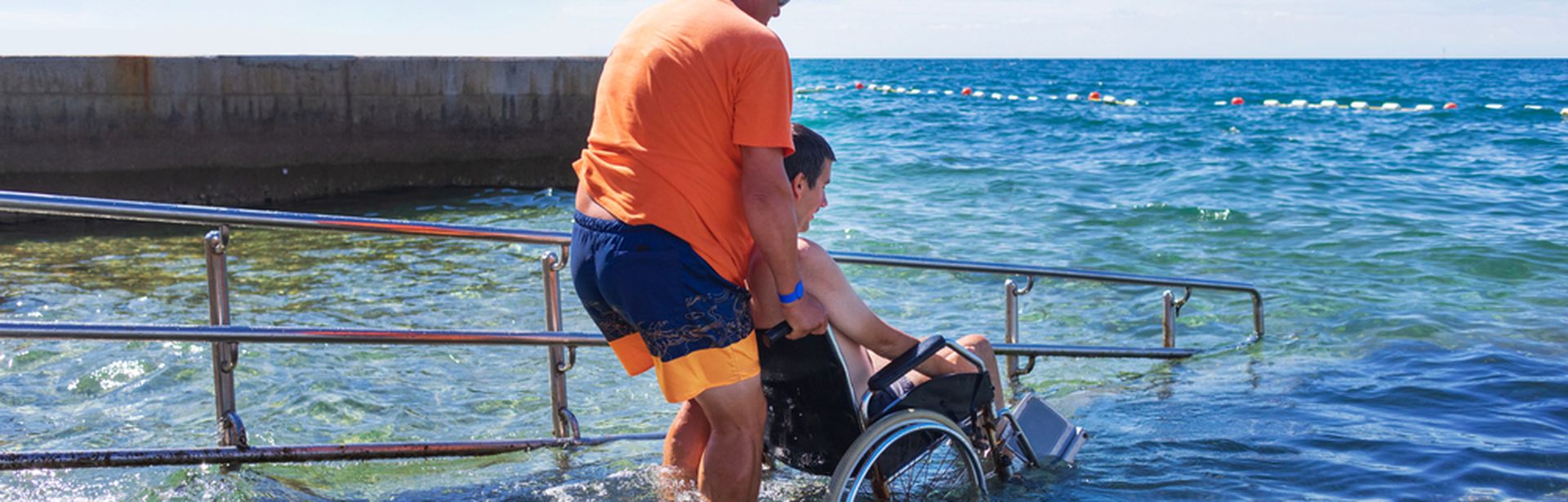Mann hilft einem anderen Mann im Rollstuhl über eine Treppe ins Meer