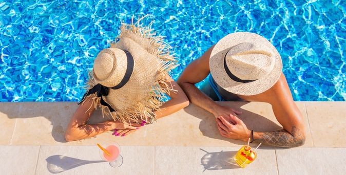 Zwei Personen von oben fotografiert, Sie tragen Strohhüte und man sieht Ihre Gesichter nicht. Sie sind in einem Pool und lehnen am Rand, vor Ihnen stehen Getränke auf dem Boden