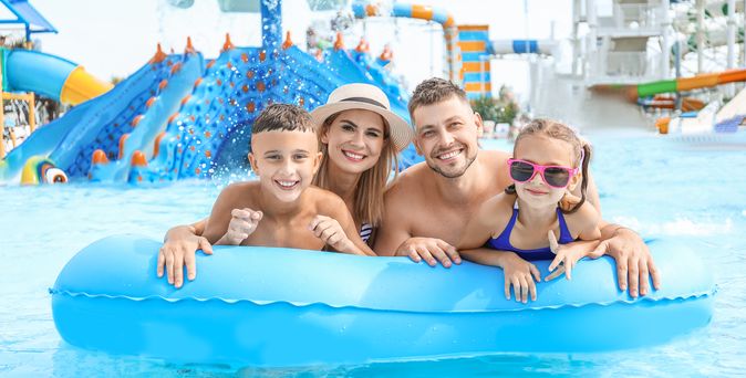 Eine Familie auf einem blauen Pool Floatie lächelt in die Kamera, im Hintergrund ist ein Aquapark