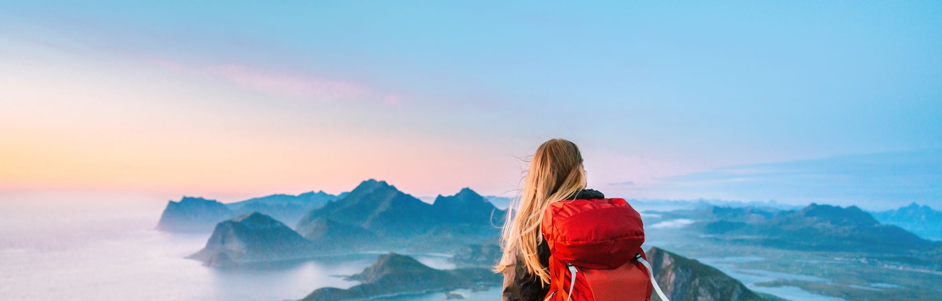 Eine Frau von hinten, Sie trägt einen großen Rucksack und schaut von einem Berg aus den Sonnenaufgang an