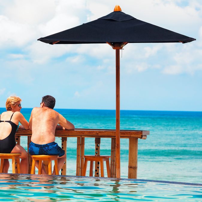 Eine Frau und ein Mann in Badekleidung sitzen an einer Holztheke auf Holzstühlen vor einem Pool und schauen auf das Meer