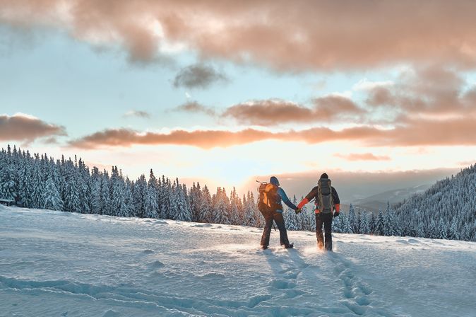 Zwei Personen stehen auf einem Schneebedeckten Berg und schauen Händchen haltend in den Sonnenuntergang