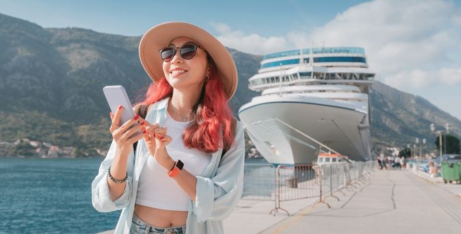 Eine Frau mit roten Haaren steht an einem Hafen, hält ihr Smartphone fest und lächelt; Im Hintergrund steht ein Kreuzfahrtschiff