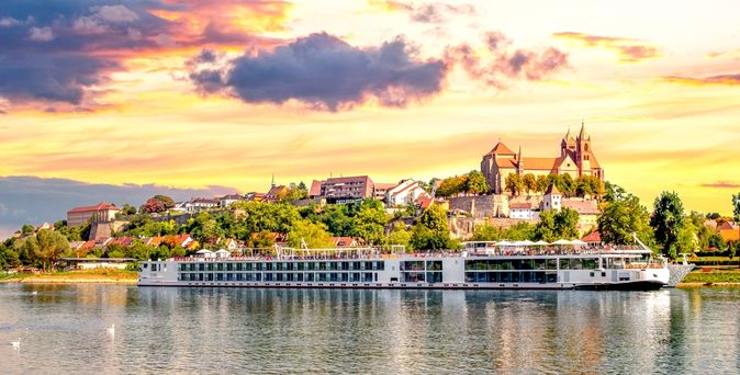 Langes Flusskreuzfahrschiff fährt entlang eines kleinen Dorfes, bei gelb leuchtendem Himmel