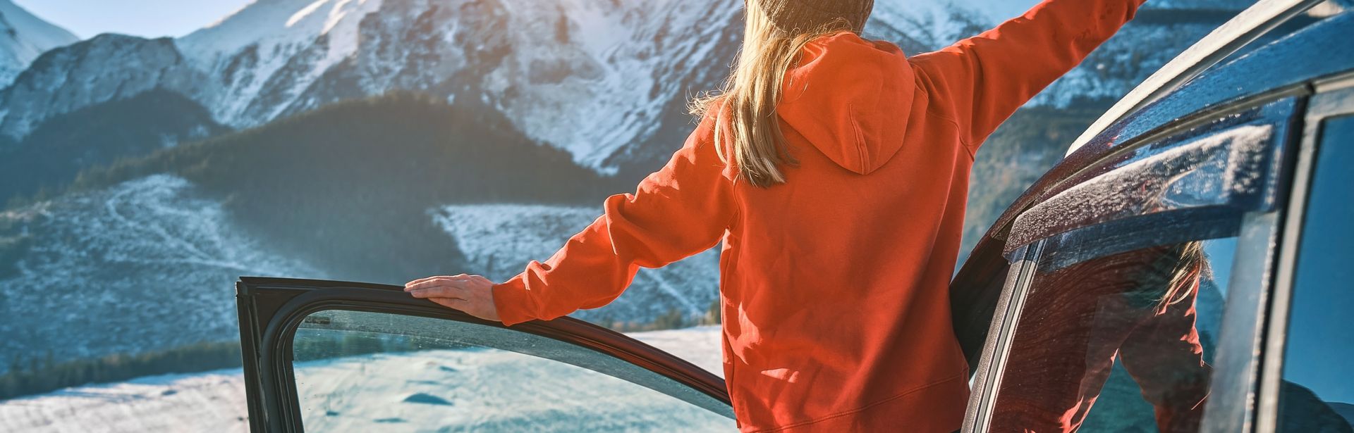 Eine Frau lehnt aus der offenen Autotür und schaut auf Schneebedeckte Berge