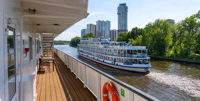 Reling eines Flusskreuzfahrtschiffes, von der aus ein weiteres Schiff zu sehen ist, mit Hochhäusern im Hintergrund