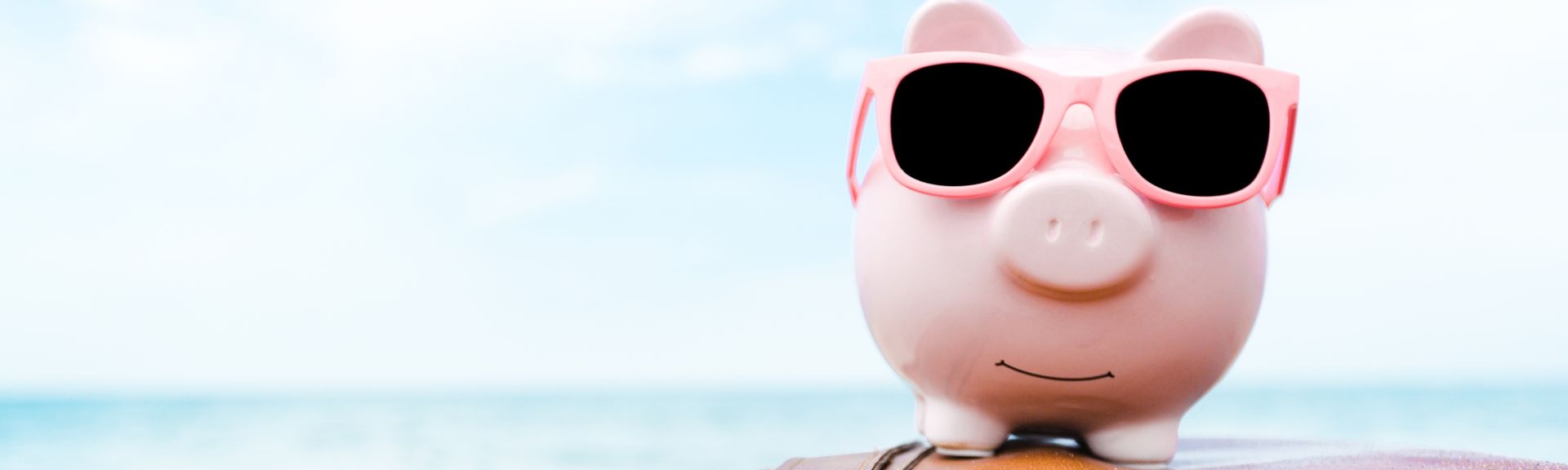 Ein rosanes Sparschwein mit Sonnenbrille sitzt auf einem Koffer am Strand
