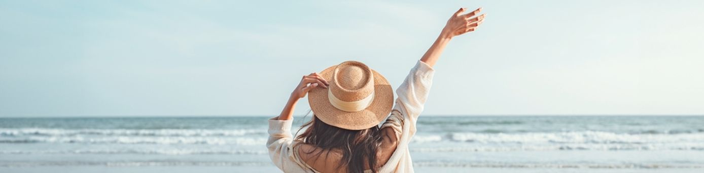 Eine Frau mit Hut von hinten, Sie schaut vom Strand aus auf das Meer und hebt den Arm 