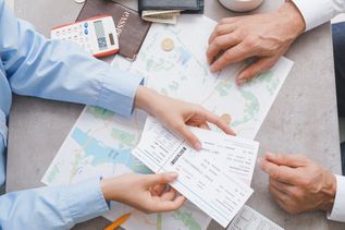 Auf einem Tisch liegen eine Landkarte, ein Taschenrechner, ein Reisepass und eine Geldbörse. Zwei Paar Hände sind zu sehen, ein Paar Hände hält Flugtickets