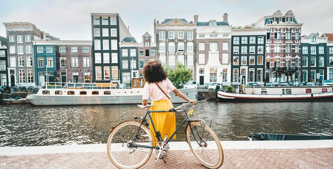 Eine Frau steht mit ihrem Fahrrad vor der Amstel und schaut auf die Häuser auf der gegenüberliegenden Seite