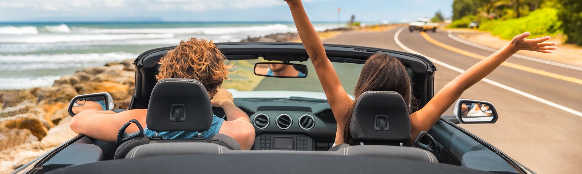 Zwei Personen im Auto mit offenem Verdeck, im Hintergrund kann man das Meer sehen