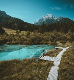 Eine Große Wiese mit einem Holzweg, auf einem Steg steht eine Frau und breitet die Arme aus, sie schaut auf den See vor ihr