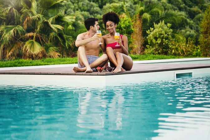 Paar sitzt mit Cocktails in der Hand und Obstschälchen auf der Terrasse vor einem Pool