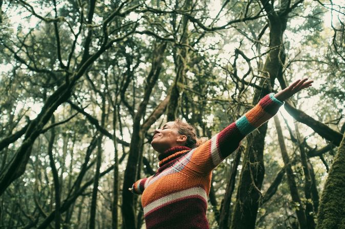 Eine Frau breitet glücklich die Arme aus und hat die Augen geschlossen, Sie steht in einem Wald voller dichter Bäume