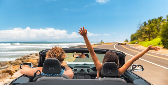Zwei Personen im Auto mit offenem Verdeck, im Hintergrund kann man das Meer sehen