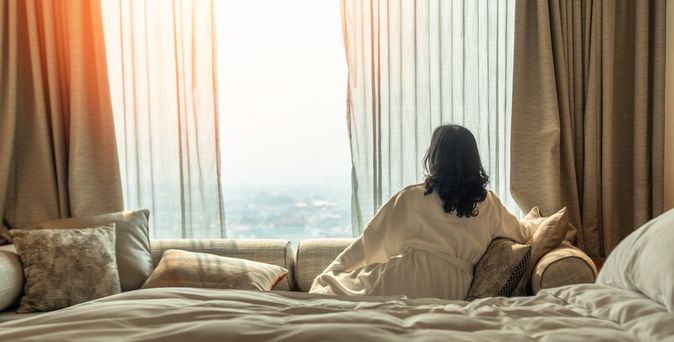 Eine Frau im Bademantel sitzt in einem Hotelzimmer auf dem Sofa und schaut aus den Fenster