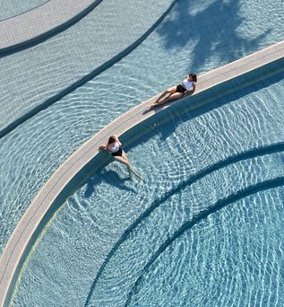 Zwei Frauen an zwei riesigen Pools; Eine liegt im Pool, während die andere sich auf der Trennung beider Pools sonnt