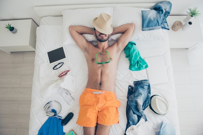 Ein Mann in oranger Badehose liegt auf dem Rücken im Bett, sein Hut liegt auf seinem Gesicht und um ihn herum liegen Anziehsachen, ein Tablet und eine Sonnenbrille