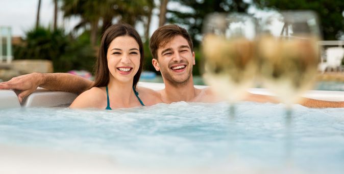 Eine Frau und ein Mann sitzen in lachend in einem Whirlpool, verschwommen sieht man im Vordergrund Weingläser
