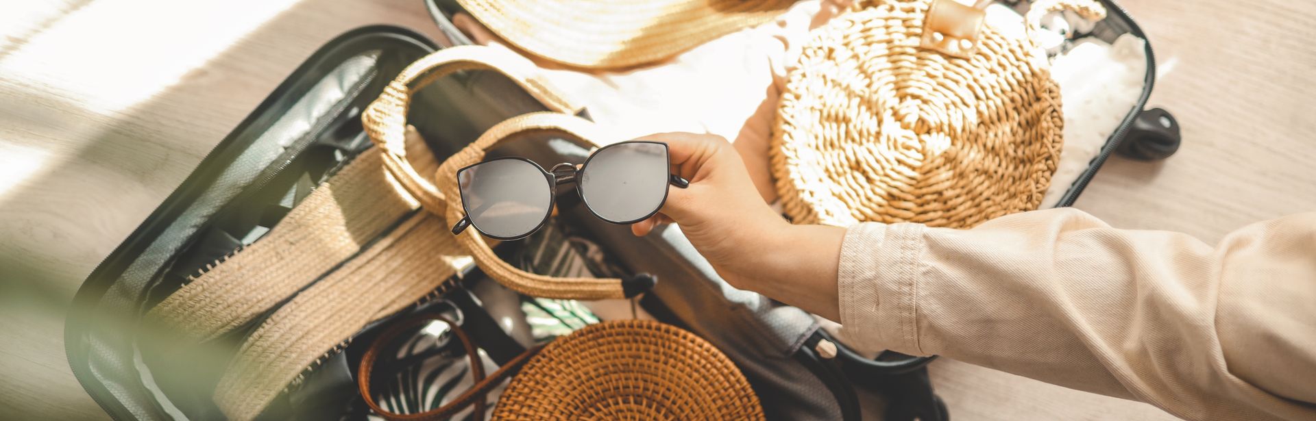 Ein offener Koffer, eine Hand legt eine Sonnenbrille zu den bereits gepackten Dingen