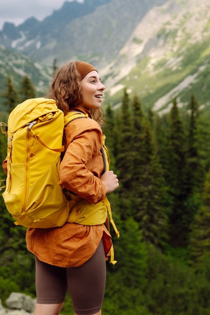 Eine Frau mit gelbem Wanderrucksack steht auf einem Berg und schaut begeistert in die Ferne