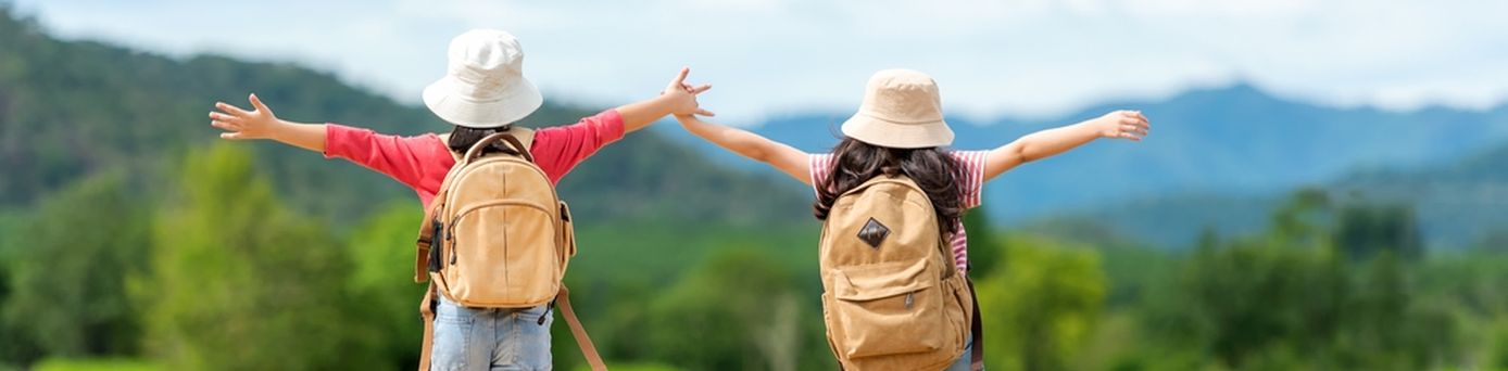 Zwei Mädchen mit Rucksäcken laufen fröhlich einen Weg entlang und heben die Arme in die Luft