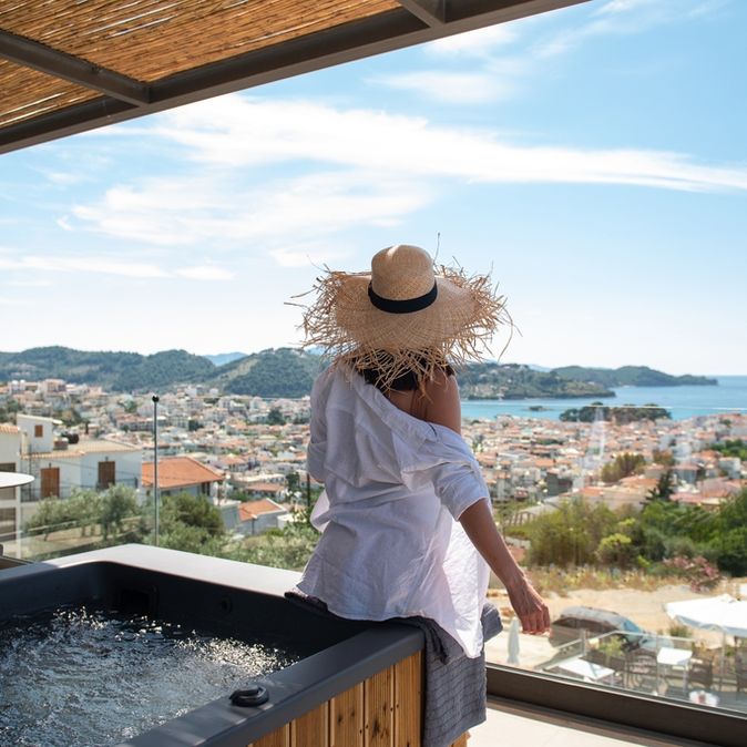 Eine Frau mit Sonnenhut sitzt auf dem Rand eines Whirlpools auf einem Balkon und blickt runter Richtung der Stadt