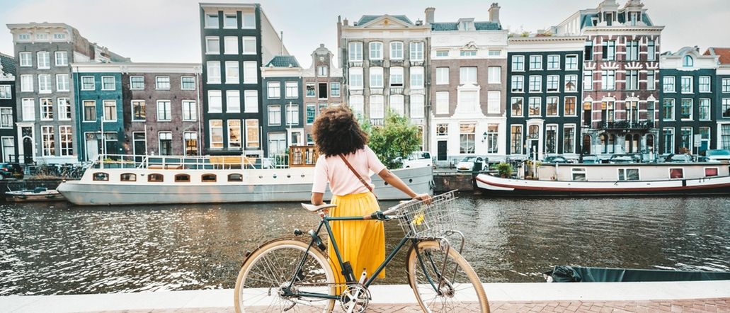 Eine Frau in einem gelben Rock steht an ihrem Fahrrad und schaut auf die Amstel und die gegenüberliegenden Häuser und Boote in Amsterdam