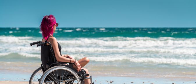 Frau im Rollstuhl mit einem Bein sitzt entspannt am Strand und blickt aufs Meer