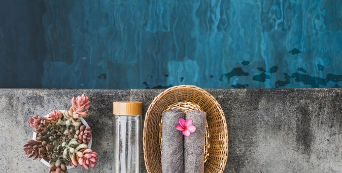 Ein Teil eines Pools aus der Vogelperspektive, ein Korb mit Handtüchern, eine Glasflasche und eine Pflanze im Topf stehen am Rand