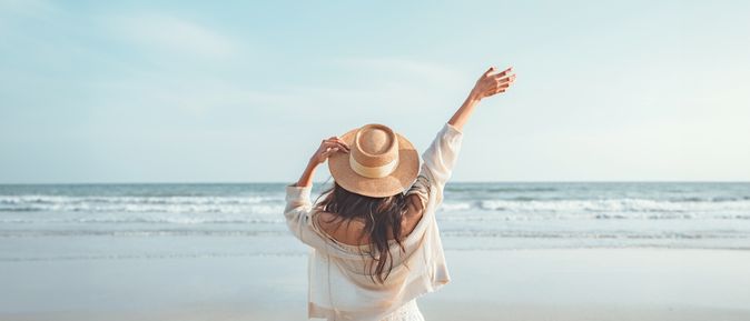 Eine Frau mit Hut von hinten, Sie schaut vom Strand aus auf das Meer und hebt den Arm 