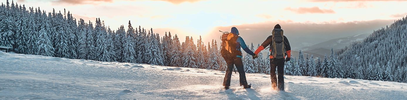 Zwei Personen stehen auf einem Schneebedeckten Berg und schauen Händchen haltend in den Sonnenuntergang