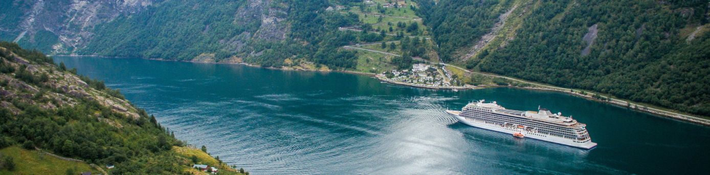 Ein Kreuzfahrtschiff fährt durch einen tiefen, von grünen Bergen gesäumten Fjord mit einem kleinen Dorf im Vordergrund.
