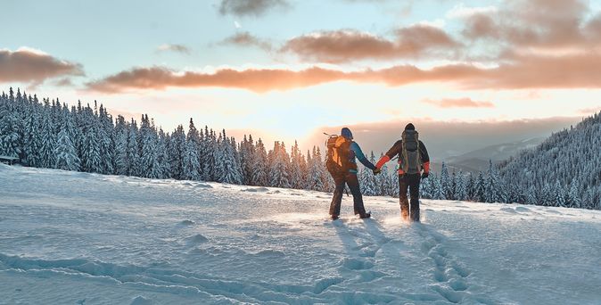 Zwei Personen stehen auf einem Schneebedeckten Berg und schauen Händchen haltend in den Sonnenuntergang