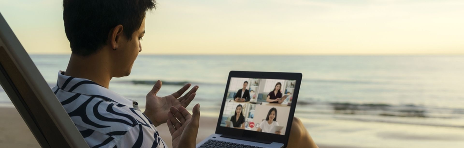 Ein Mann sitzt in einem Liegestuhl am Meer mit Laptop auf dem Schoß und nimmt an einer Videokonferenz teil