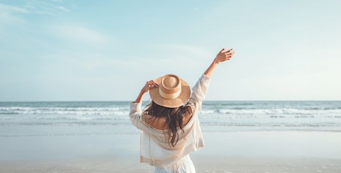 Eine Frau mit Hut von hinten, Sie schaut vom Strand aus auf das Meer und hebt den Arm 