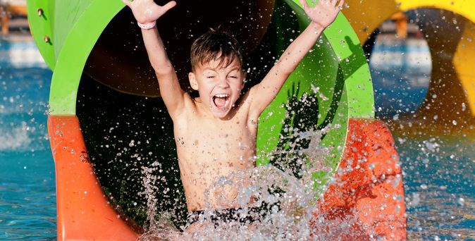 Ein Junge rutscht aus einer Wasserrutsche in einen Pool und reißt die Arme hoch