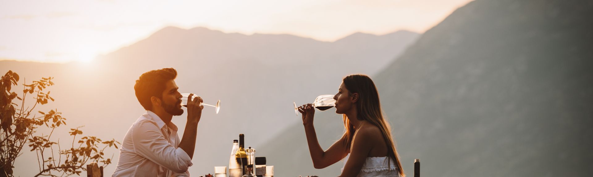 Eine Frau und ein Mann sitzen sich bei Sonnenuntergang an einem Tisch am Wasser gegenüber und trinken Wein