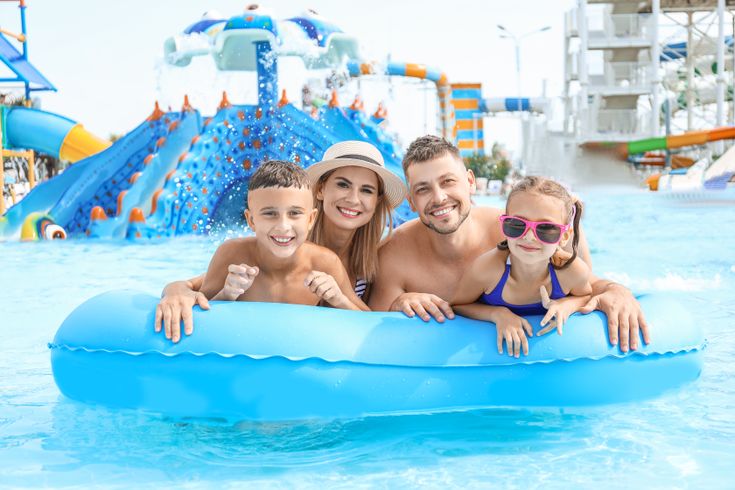 Eine Familie auf einem blauen Pool Floatie lächelt in die Kamera, im Hintergrund ist ein Aquapark
