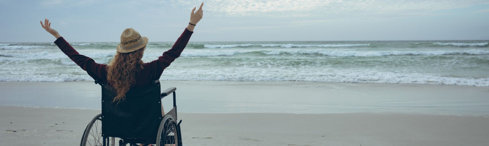 Frau im Rollstuhl hebt die Arme und schaut aufs Meer