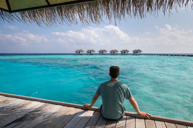 Ein Mann sitzt auf einem Steg am Meer und schaut auf mehrere Wasserbungalows