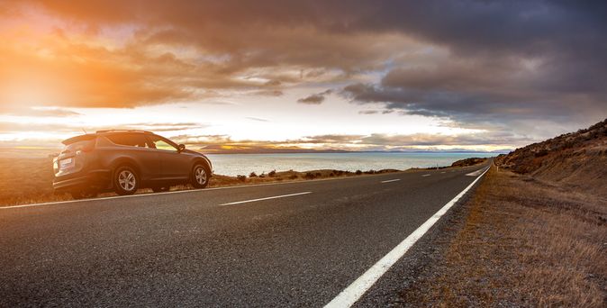Ein Auto fährt bei Sonnenaufgang an der Kamera vorbei auf einer Straße am Wasser.