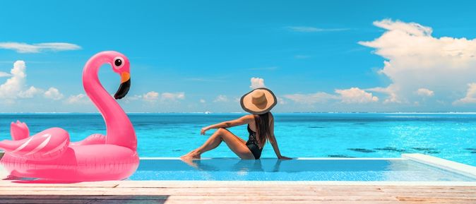 Frau sitzt am Rand eines Pools mit Meerblick, neben Ihr ein aufblasbarer Flamingo