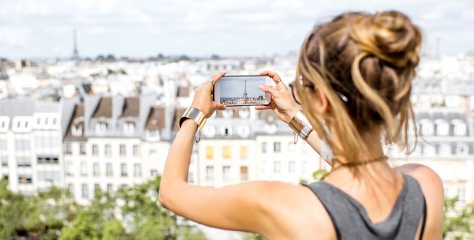 Eine Frau macht mit ihrem Handy ein Foto von einer Stadt, welche sie von einem höher gelegenen Aussichtspunkt betrachtet