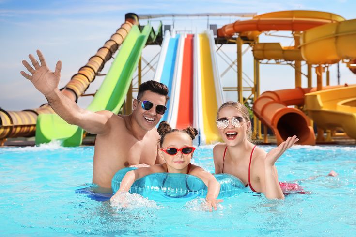 Eltern und ein Kind in einem Pool, Das Kind trägt eine Sonnenbrille und schwimmt in einem Schwimmring, im Hintergrund sind Wasserrutschen
