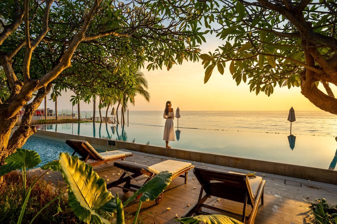 Frau steht bei Sonnenuntergang vor einem Pool der direkt vor einem Meer liegt