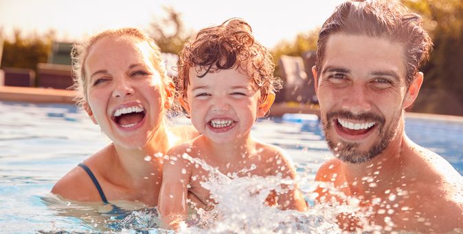 Eltern mit einem Kind spielen im Pool und lachen in die Kamera