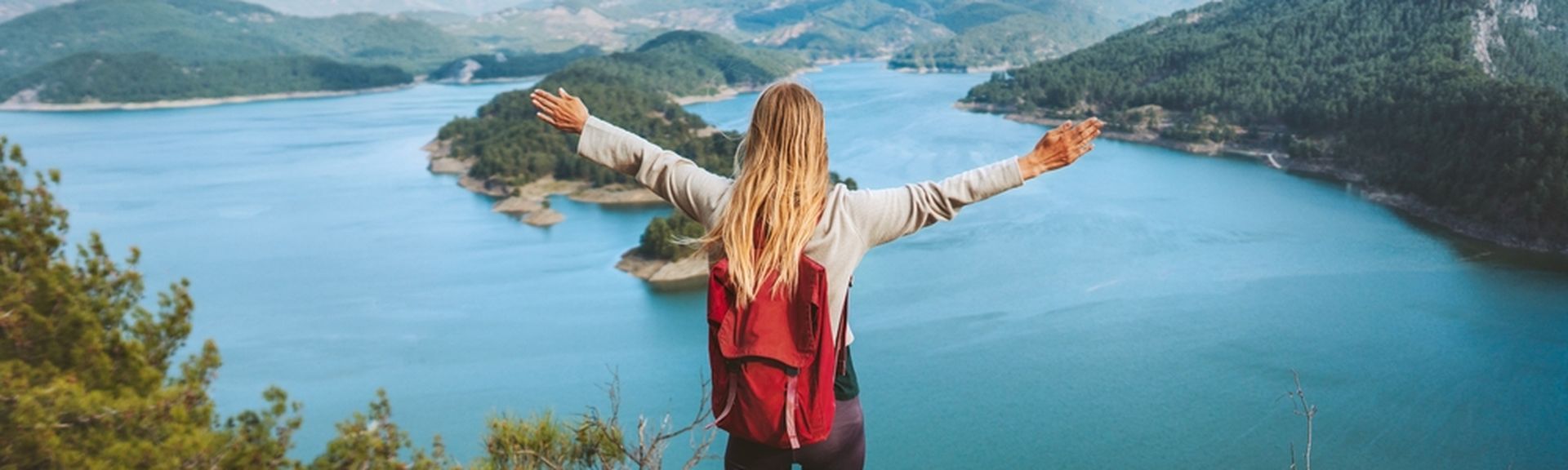 Eine Frau mit Rucksack schaut von einer Klippe auf einen großen See, eine Berglandschaft und breitet die Arme aus